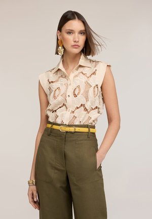 Młoda kobieta o fryzurze bob, ubrana w bezrękawowy, beżowy koronkowy top, oliwkowe szerokie spodnie, żółty pasek, złote kolczyki i bransoletki, z ręką w kieszeni.