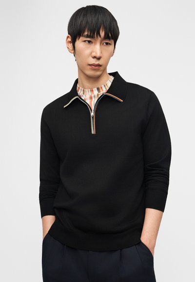 Νεαρός άνδρας με μαύρο μακρυμάνικο πουλόβερ με μισό φερμουάρ και ριγέ γιακά πάνω από πολύχρωμο πουκάμισο με ρίγες, με τα χέρια στις τσέπες του σκούρου μπλε παντελονιού.