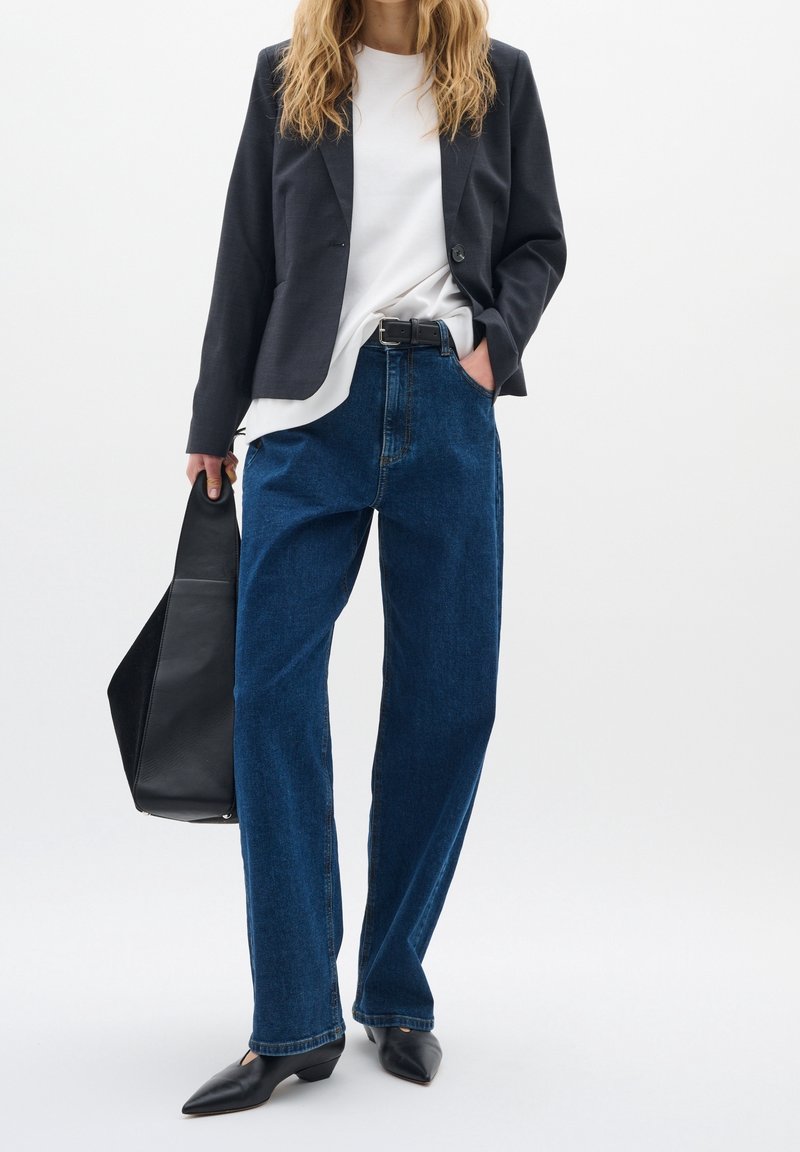 Femme portant un blazer noir, une chemise blanche, un jean bleu et des chaussures noires, tenant un grand sac à main noir dans une main.