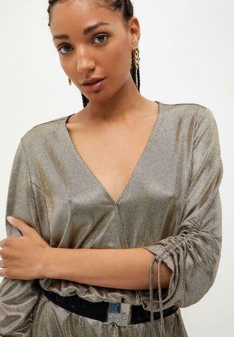 Metallic bronzen jurk met een diepe V-hals en lange mouwen, met geschuurde accenten bij de mouwen en een zwarte riem met een zilveren gesp.