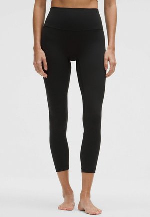 Kobieta ubrana w czarne, krótkie legginsy z wysokim stanem, stojąca boso na jednolitym szarym tle.