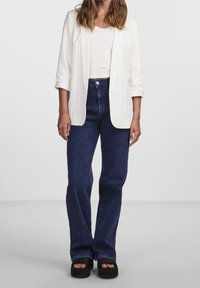 Blazer blanc sur un haut crème, associé à un jean bleu foncé taille haute. Des sandales à plateforme noires complètent l'ensemble.