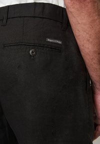 Detailní záběr na zadní kapsu černých lněných kalhot s knoflíkem a viditelnou značkou Marc O’Polo, vedle paže osoby se světlou pletí a vlasy.