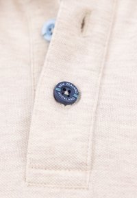 Beige poloshirt met een blauwe knoop en de tekst "NEW ZEALAND AUCKLAND" en een doorgestikte knoopsgat. Geverfd katoen met een textuur.