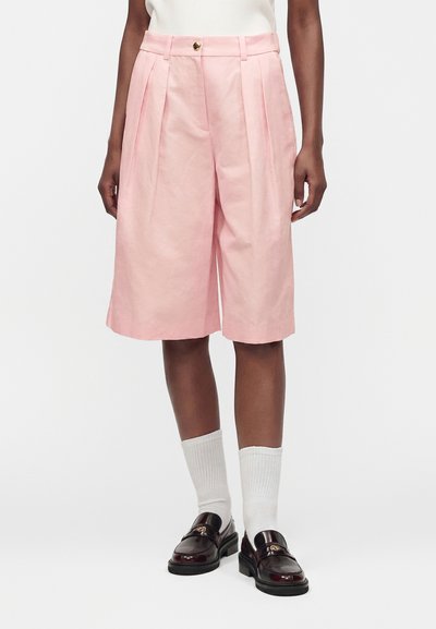 Personne portant un short plissé rose taille haute longueur genou avec des chaussettes blanches et des mocassins en cuir marron foncé sur fond blanc.