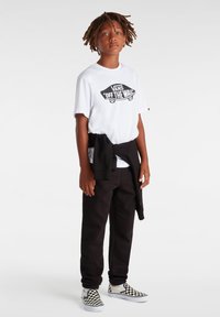Witte t-shirt met zwart "VANS OFF THE WALL" logo, zwarte joggingbroek en zwart-wit geruite instap schoenen, met een sweatshirt om de taille geknoopt.
