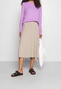 Lila Pullover mit gerippter Struktur, beige gefalteter Midirock und schwarze Slipper-Sandalen. Weiße Tote-Bag in der Hand gehalten.