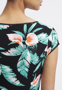 Kobieta z odsłoniętym ramieniem i szyją, nosząca czarną bluzkę w dużą zieloną roślinność oraz różowo-pomarańczowy wzór kwiatowy.