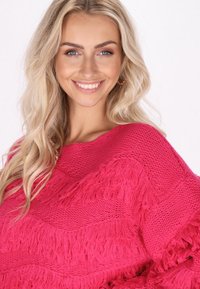 Rosa stickad tröja med texturerad frans, vid halslinning och avslappnad passform. Designen inkluderar horisontella ränder med varierande stickmönster.