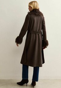 Abrigo marrón con cuello y puños de piel sintética, ceñido en la cintura. Textura suave, hasta la rodilla, combinado con jeans oscuros y botas de tacón.