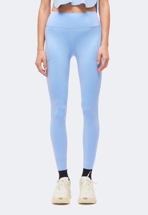 Kvinde iført lyseblå leggings med høj talje og sidelommer, sorte sokker og beige Nike-sneakers, stående mod en lys baggrund.