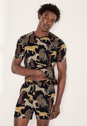 Homme portant un short noir assorti et un t-shirt avec un imprimé guépard et léopard jaune, brun et blanc, debout contre un mur blanc.