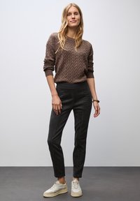 Brun texturerad tröja med ett zickzackmönster, tillsammans med svarta slim-fit byxor och ljusa sneakers, stående mot en neutral bakgrund.