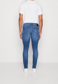 Jeans ajustados en denim azul medio con una textura suave, que presentan bolsillos traseros y una cinturilla negra. Se llevan con una camiseta blanca lisa y zapatillas.