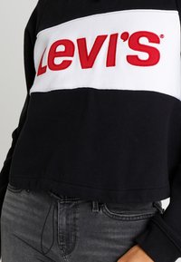 Felpa nera corta con una banda orizzontale bianca che presenta il logo "Levi's" in rosso in lettere grasse. Tessuto misto cotone con una superficie liscia.