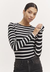 Langärmliges gestreiftes Oberteil; schwarz-weiße horizontale Streifen; weicher Stoff; Rundhalsausschnitt; enganliegendes Design mit verlängerten Bündchen; kombiniert mit schwarzen Jeans.
