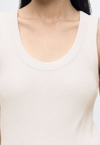 Femme portant un haut sans manches, côtelé, beige clair avec un col rond, laissant apparaître son cou et ses clavicules.