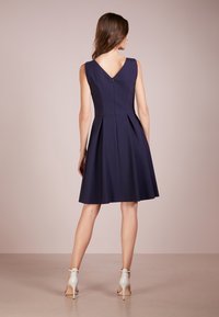 Marineblaues A-Linien-Kleid mit V-Ausschnitt, tailliertem Oberteil und plissiertem Rock. Hat einen Reißverschluss und einen weichen Stoff. Getragen mit weißen High Heels.