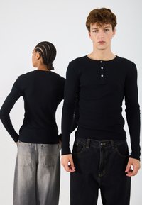 Črna dolga majica z dolgimi rokavi in okroglim izrezom, z zapenjanjem na tri gumbe, kombinirana z visokim pasom, temno in svetlo sivo denim hlačami.