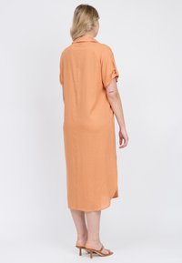 Vestido de manga corta y cuello en tela naranja tenue; presenta un corte relajado, aberturas laterales y un detalle de botones en la parte posterior.