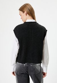 Schwarz gestrickte Weste mit Zopfmuster, kurzen Ärmeln und Rippenbund, getragen über einem weißen Langarmshirt. Rückansicht, lässiges Design.