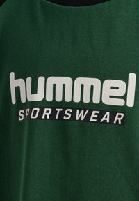 Grön sporttröja med tydlig vit 'hummel' text och en svart rektangulär 'SPORTSWEAR' detalj. Mjuk tygtextur, rundad halsdesign.