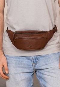 Uomo che indossa jeans azzurri chiari e una camicia bianca con una cintura a tracolla in pelle marrone, chiusa e fissata intorno alla vita.