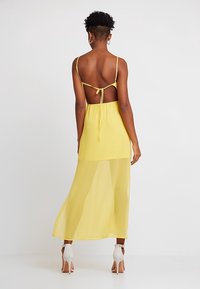 Robe en mousseline jaune avec un design noué dans le dos et des fines bretelles ; tissu transparent s'étendant jusqu'aux chevilles, assortie à des talons hauts blancs.