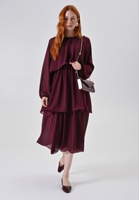 Abito in chiffon borgogna a strati con maniche lunghe e un design fluido. Abbinato a una borsa marrone e mocassini lucidi coordinati.