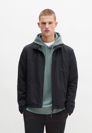 Jonge man met kort blond haar, die een zwart vest met ritssluiting draagt over een groene hoodie en zwarte broek, staand met handen in de zakken.