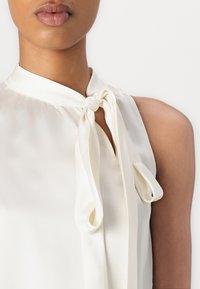 Blouse en soie de couleur blanc cassé avec un détail à nouer au niveau du col, présentant une texture légèrement froncée et une finition lisse. Modèle sans manches.