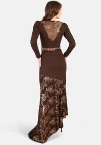 Robe longue marron avec manches longues, dotée d'un dos transparent et d'une broderie florale. La jupe présente un design fluide avec un détail de traîne.