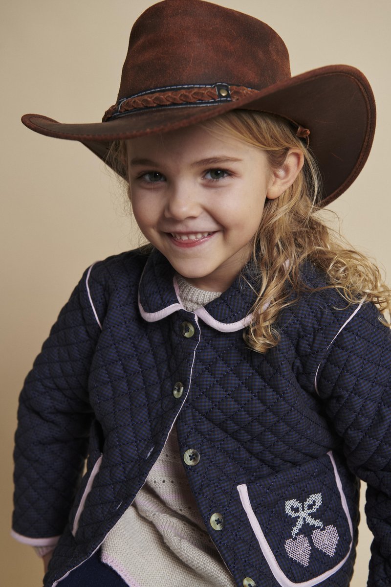 Tamsiai mėlyna dygsniuota striukė su rožiniu apvadu, puošniomis kišenėmis su siuvinėtais širdelių ir lankų motyvais. Modelis dėvi rudą kaubojišką skrybėlę.