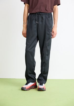 Personne portant un pantalon noir à cordon avec un petit logo sur la cuisse, assorti à des baskets dégradées blanc, rouge et noir sur un sol vert.