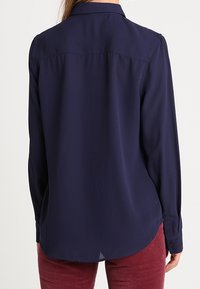 Navy blue long-sleeve shirt with a collar, smooth texture, button cuffs, and rounded hem. Features a yoke with subtle stitching detail.