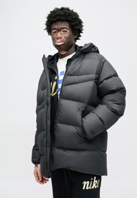 Czarna kurtka puchowa z kapturem, pikowana, z zamkiem z przodu, noszona na białej koszuli z niebieskimi i złotymi akcentami oraz czarnych spodniach dresowych.