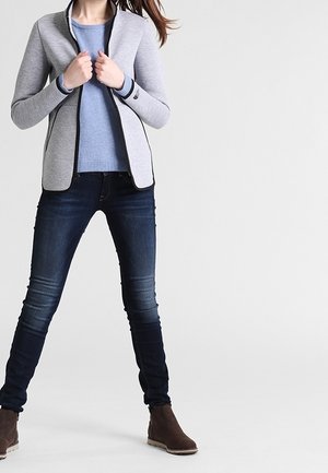 Nainen pukeutuneena vaaleanharmaaseen vetoketjulliseen takkiin sinisen neuleen päällä, tummiin kapealahkeisiin farkkuihin ja ruskeisiin nilkkureihin, seisoo jalat hieman erillään.