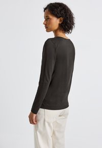 Haut à manches longues, vert foncé, en tissu doux avec un décolleté rond, des manches ajustées et une coupe droite. Porté avec un pantalon de couleur claire.