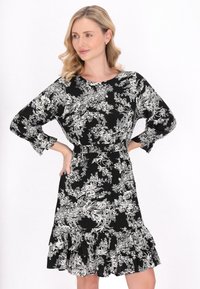 Schwarzes Blumenkleid mit weißen botanischen Mustern, rundem Ausschnitt, langen Ärmeln und einem ausgestellten, geflammten Saum. Leichter Stoff.
