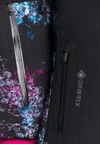 Gros plan sur deux vestes, l'une avec un motif abstrait coloré et une fermeture éclair étanche, l'autre noire avec le logo Gore-Tex et une poche zippée verticale.