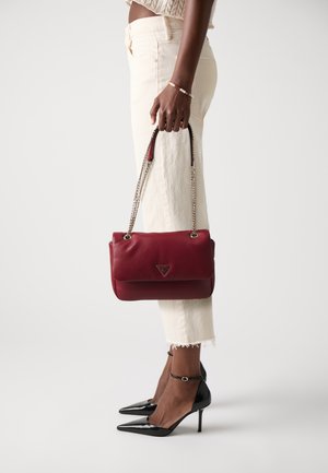 Donna che indossa pantaloni color crema e tacchi neri, mentre tiene una borsa a tracolla rossa in pelle con una catena dorata.