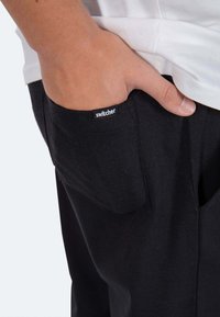 Schwarze Hose mit einer Seitenasche, die ein kleines "Switcher"-Etikett hat. Der Stoff wirkt weich und dehnbar und hebt ein einfaches, praktisches Design hervor.