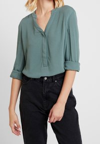 Frau in einer hochgekrempelten teal-blauen Bluse, die in hochtaillierte schwarze Jeans gesteckt ist, vor einem schlichten Hintergrund.