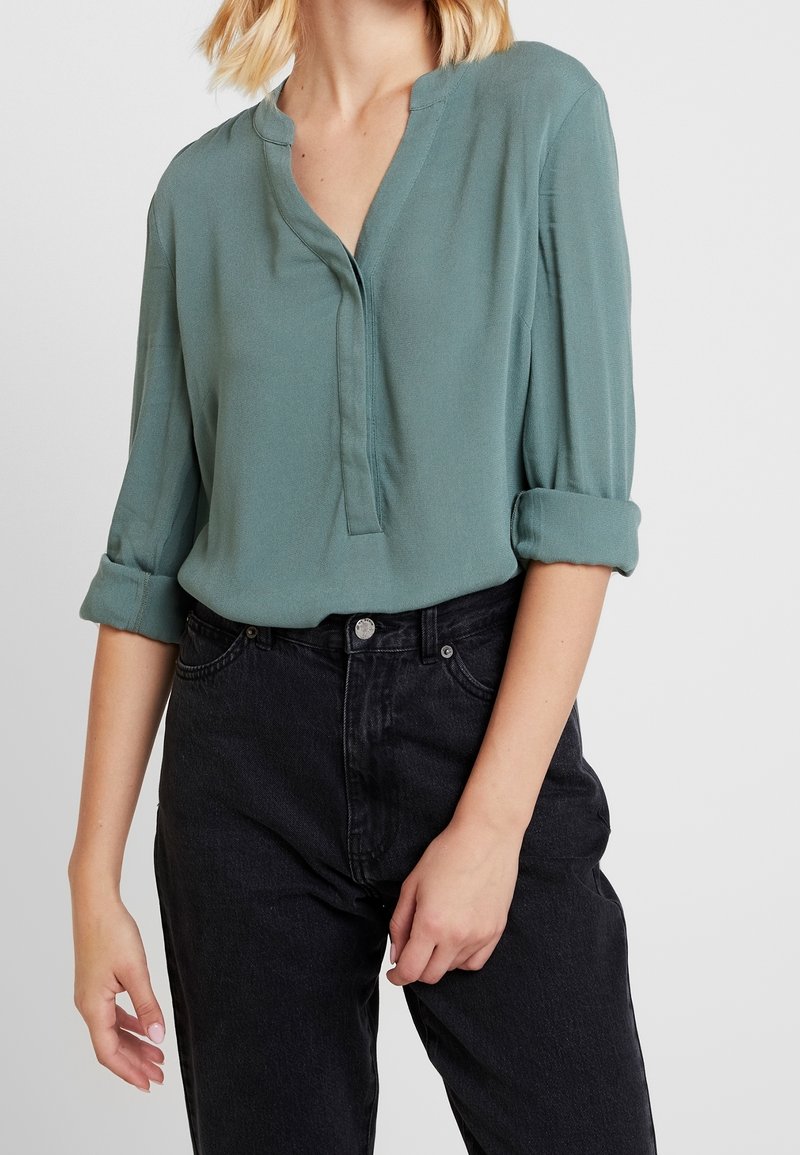 Frau in einer hochgekrempelten teal-blauen Bluse, die in hochtaillierte schwarze Jeans gesteckt ist, vor einem schlichten Hintergrund.