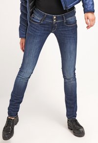 Blue denim skinny jeans with subtle fading, classic five-pocket design, two-button closure, and topstitching details. Worn with black shoes.