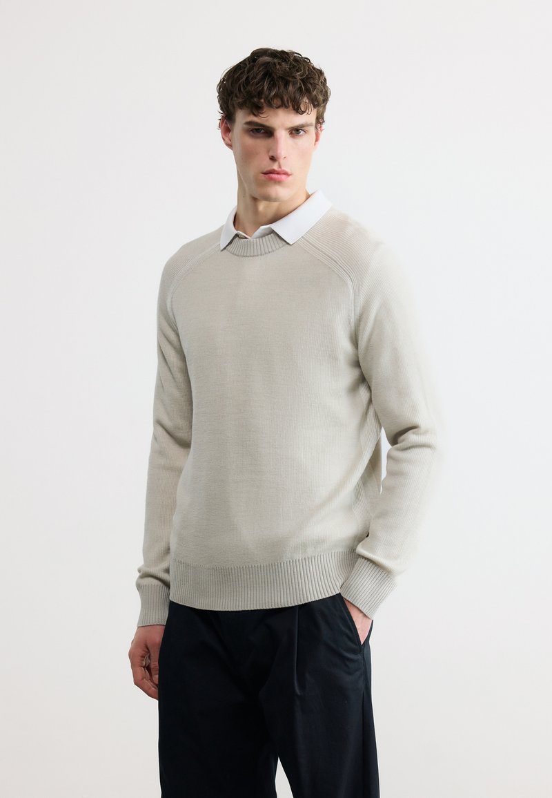 Jeune homme aux cheveux courts et bouclés portant un pull beige clair sur une chemise blanche à col et un pantalon foncé, debout devant un fond blanc uni.