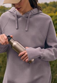 Sweatshirt gris à coupe décontractée, avec une capuche à cordon de serrage et des poignets côtelés, tenu par des mains serrant une bouteille d'eau en acier inoxydable.