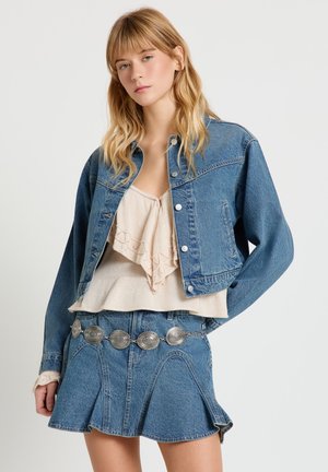 Jeune femme portant une veste en jean bleue et une jupe en jean évasée avec une ceinture argentée à conchos et un haut beige superposé.