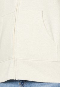 Haut à capuche de couleur crème en tissu doux, doté d'une poche avant et d'un ourlet côtelé. Le design comprend des détails de couture visibles.