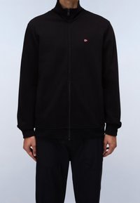 Schwarze Zip-Jacke mit hohem Kragen, aus weichem Stoff gefertigt. Verfügt über ein kleines rotes Logo auf der linken Brust. Gerippte Bündchen und Saum.